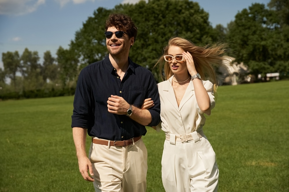 A beautiful young couple in elegant clothing stroll through a lush green field in a display of old-money style and rich people lifestyle.