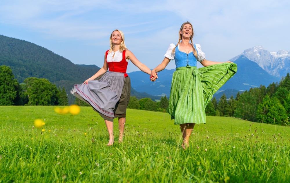 Two joyful women in traditional Bavarian dresses, dirndl or tracht holding hands and twirling in a meadow with mountains behind