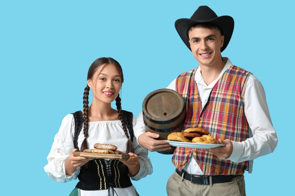 Young couple in traditional German clothes with barrel of beer, sausages and pretzels on blue background.