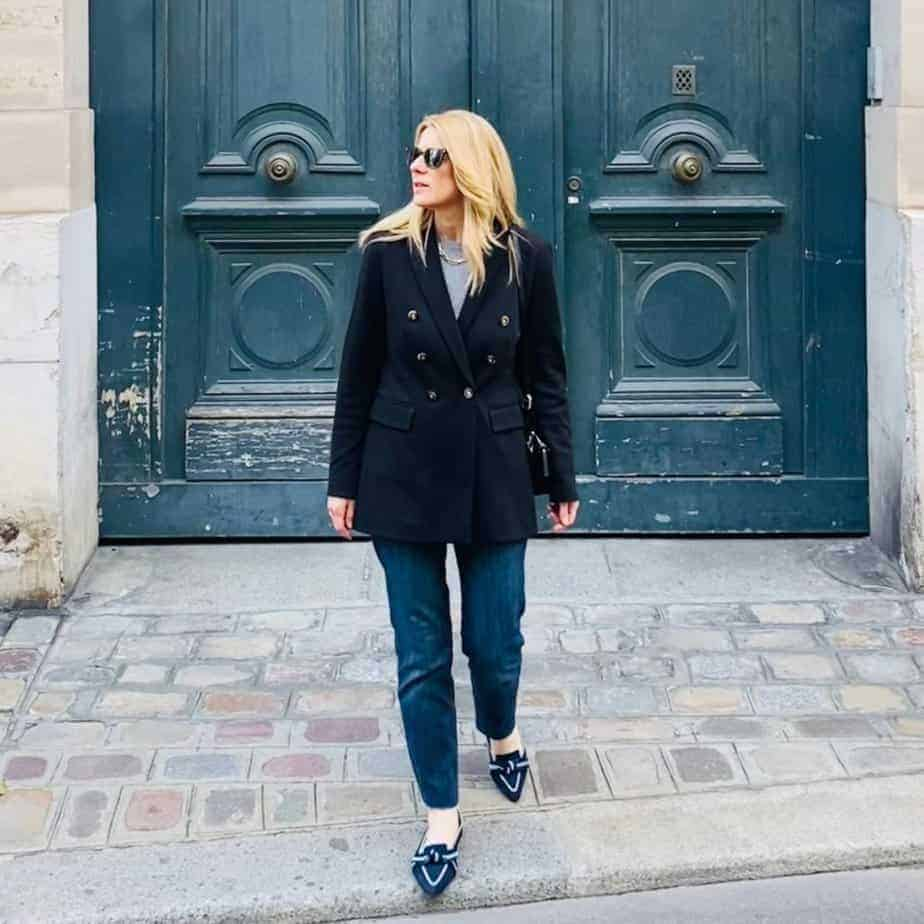 a woman wearing well fitted pants loafers and glasses standing infront of blue wooden door