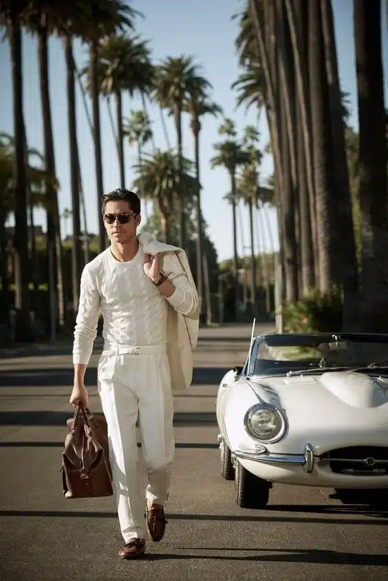 boy walking on a road wearing white pants weaters and blazor holding a brown bag